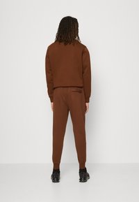 Osoba s dredy oblečená v hnědém teplákovém obleku a černých teniskách, stojící čelem k jednoduché bílé zdi, s uvolněnýma rukama podél těla.