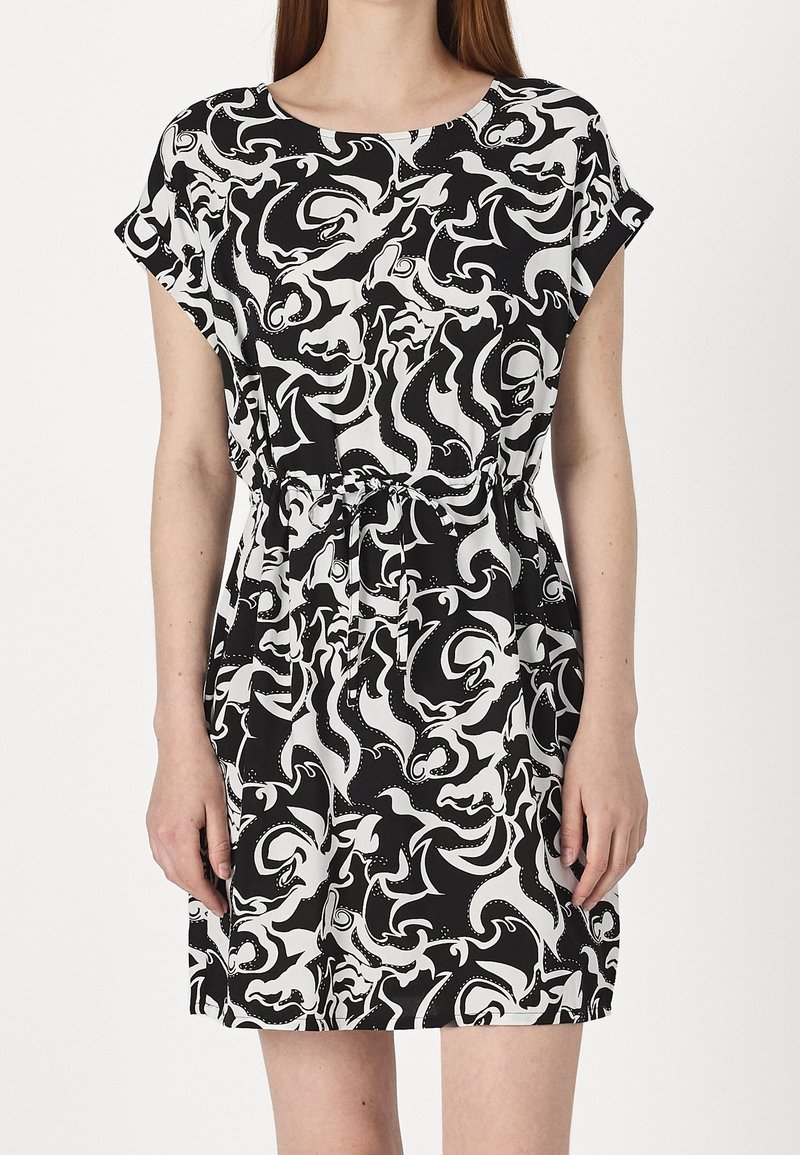 Kobieta ubrana w sukienkę z krótkim rękawem w czarno-biały abstrakcyjny wzór, z taliowaniem i okrągłym dekoltem.