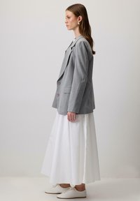 Pilkas švarkas su smulkiomis juostelėmis, turintis didelius atvartus, du priekiniai mygtukai ir šoninės kišenės, dėvimas ant baltos, laisvai krentančios sijono ir lengvų batų.