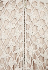 Superposition de tissu en dentelle avec un motif floral en beige clair, comportant une fermeture éclair centrale. L'arrière-plan est en matériau beige uni. Détails texturés évidents.