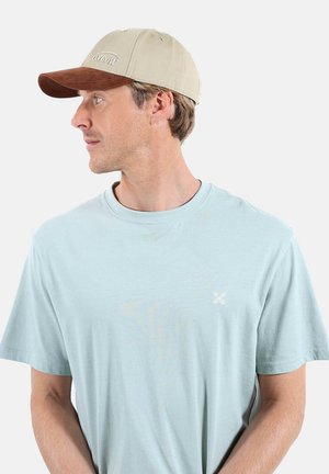 Homme portant un t-shirt bleu clair et une casquette beige à visière marron, regardant vers la gauche sur un fond blanc uni.