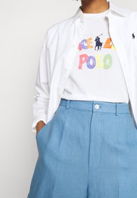 Vit skjorta med knappar över en vit t-shirt med färgglad "Polo"-text, tillsammans med ljusblå linbyxor med sidofickor.