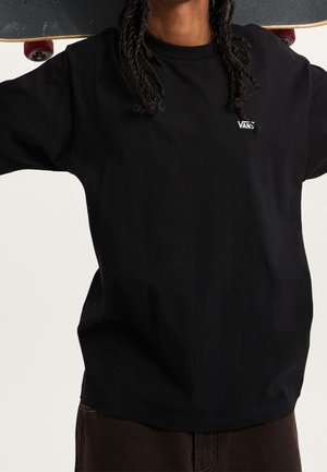 Personne portant un t-shirt Vans noir tenant un skateboard derrière son cou avec les deux mains, visible du cou à la taille.