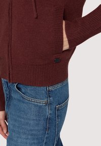 Mörkröd stickad cardigan med ribbade ärmar och nederkant, med en framficka. Kombinerad med blå denimjeans med en klassisk passform.