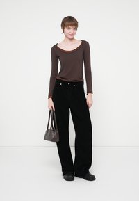 Maglietta marrone a maniche lunghe con collo a contrasto, abbinata a pantaloni in corduroy neri a gamba larga. Borsa marrone, scarpe nere, sfondo semplice.