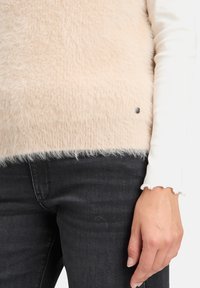 Fluffy beige trui met een zachte textuur, voorzien van een lichte kleurverloop en een zilverkleurige accentknop aan de zoom, gecombineerd met donkere jeans.
