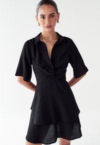 Mujer con el cabello engominado que lleva un vestido negro de manga corta con un nudo en el frente, pendientes de aro dorados y un collar delgado de oro, de pie contra un fondo blanco.