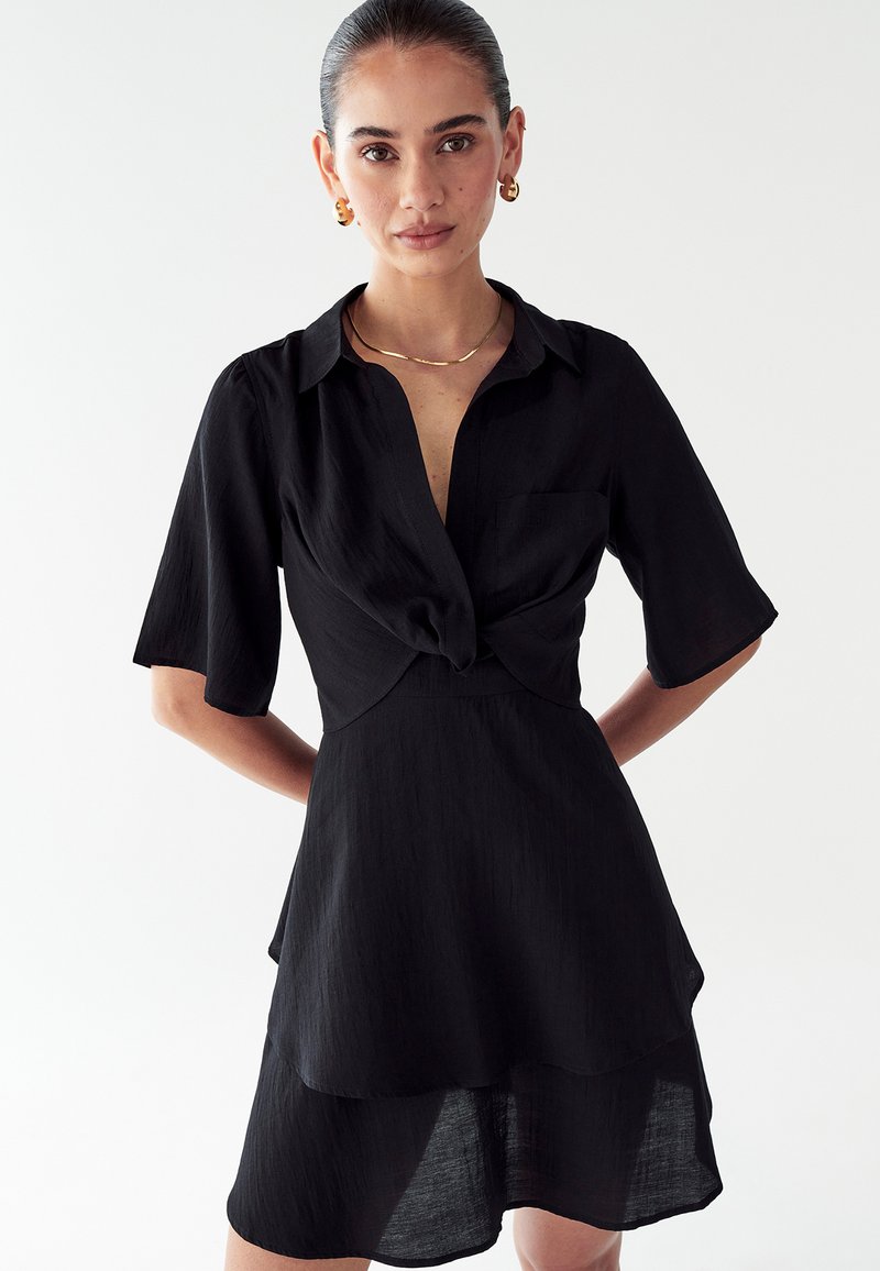 Mujer con el cabello engominado que lleva un vestido negro de manga corta con un nudo en el frente, pendientes de aro dorados y un collar delgado de oro, de pie contra un fondo blanco.