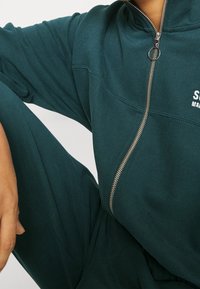 Dunkelgrüne Zip-Jacke mit hohem Kragen, silbernem Reißverschluss und weißem Logo-Druck. Der Stoff wirkt weich mit einer leicht strukturierten Oberfläche.