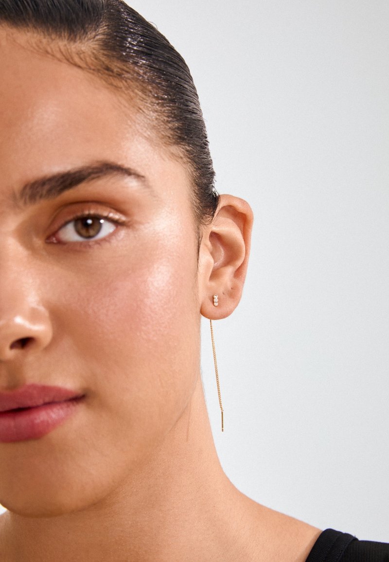 Zlatne okrugle naušnice s dugim, tankim lantanom. Jednostavan dizajn s malim akcentom blizu ušne resice. Glatka tekstura i sjajan završetak.