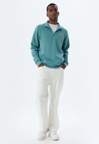 Ljusblå fleecetröja med dragkedja i kragen och en liten logotyp, matchad med vita raka byxor och vita sneakers.