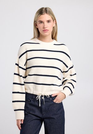 Pull crème à rayures bleu marine, texture côtelée, col rond et ourlet à cordon. Associé à un pantalon rayé sombre.