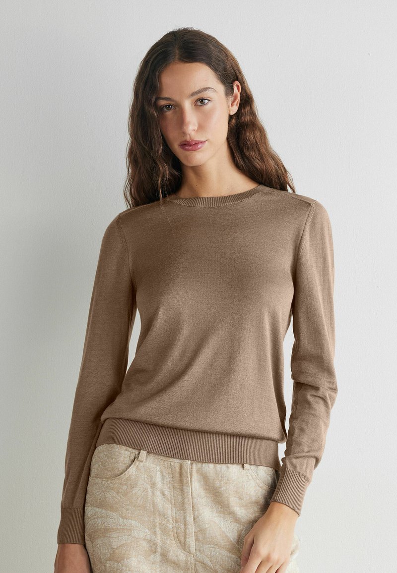 Kvinde med langt bølget hår iført en brun langærmet sweater og lyse mønstrede bukser, stående mod en ensfarvet hvid væg.