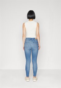 Top blanco sin mangas de forma ajustada, combinado con jeans skinny de color azul claro que tienen bolsillos traseros y una etiqueta de cuero marrón cerca de la cintura.