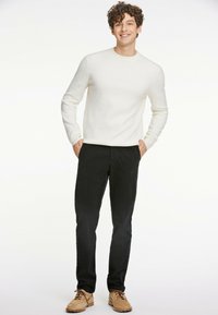 Helle beige Strickpulli mit Rundhalsausschnitt, schwarze Hose und braune Schuhe. Das Model steht mit den Händen in den Taschen und zeigt einen lässigen Schnitt und ein schlichtes Design.