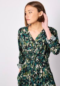 Grünes gemustertes Kleid mit V-Ausschnitt, langen Ärmeln, Knopfleiste, Taillengürtel und strukturierten Akzenten an den Bündchen.