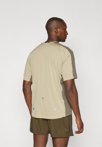 Lightweight athletic t-shirt in beige with darker green side panels, short sleeves, a rounded neckline, and reflective logo detail at the back.