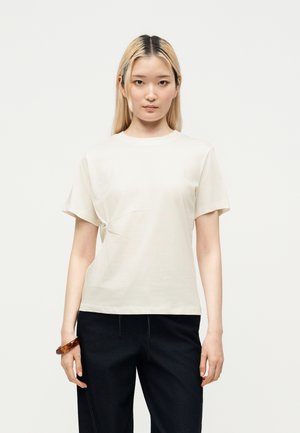 Jeune femme aux cheveux blonds et raides porte une chemise crème à manches courtes, un pantalon noir et un bracelet marron, face à un fond clair.
