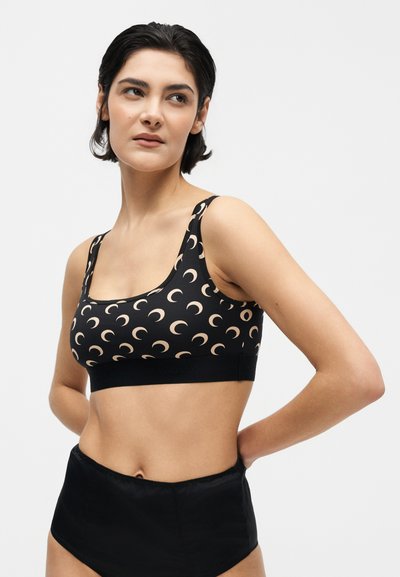 Γυναίκα με κοντά σκούρα μαλλιά φοράει μαύρο αθλητικό σουτιέν με ανοιχτό μοτίβο ημισελήνου και μαύρο κάτω μέρος με ψηλή μέση, χέρια πίσω από την πλάτη.