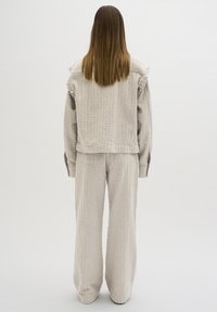 Nainen, jolla on päällään teksturoitu beige takki ja siihen sopivat leveälahkeiset housut, seisoo selkä kameraan päin tasaisella vaalealla taustalla.