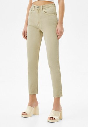 Kvinde iført beige højtaljede, tætsiddende jeans og cremefarvede mules med blokhæl i laklæder, stående foran en hvid baggrund.