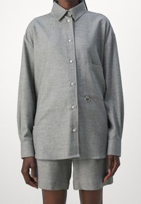 Chemise grise à boutons, avec un col, des manches longues et une poche poitrine. Elle présente un tissu texturé et une boucle décorative près de la poche.