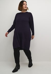 Knit sweater dress in dark purple, featuring a ribbed texture, round neckline, and drop shoulders, paired with black leggings and ankle boots.