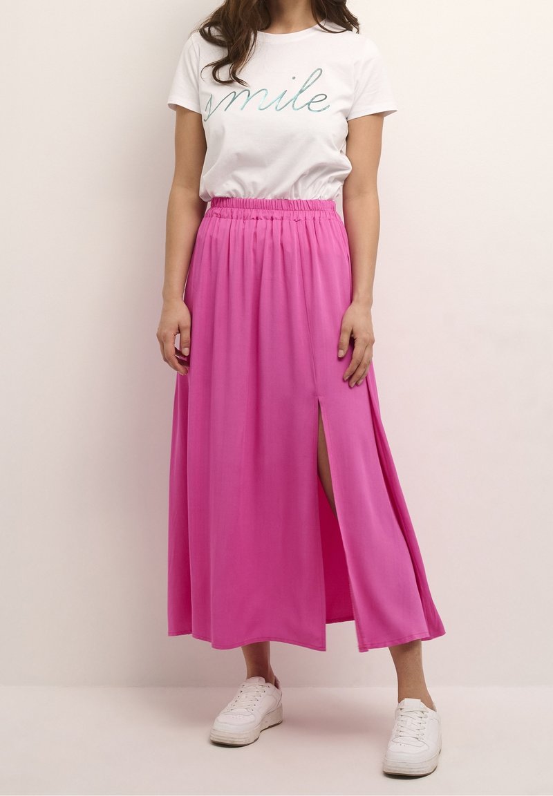 Jupe maxi rose avec une fente latérale, en tissu lisse, associée à un T-shirt blanc avec des lettres turquoise. Baskets blanches.