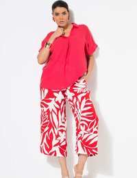 Rode korte mouwen shirt met een V-hals, gecombineerd met witte culottes met gedurfde rode tropische bladerenpatronen. Draagt witte sandalen en accessoires.