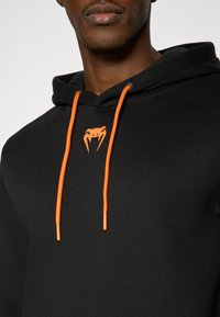 Μαύρο φούτερ με πορτοκαλί κορδόνια και λογότυπο, με απαλή υφή και κανονική εφαρμογή. Διαθέτει μίνιμαλ σχεδιασμό.