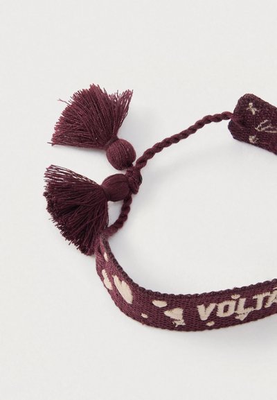 Braccialetto intrecciato color marrone scuro con due nappe, motivi a cuore e una chiusura regolabile annodata. Il materiale è un tessuto morbido.