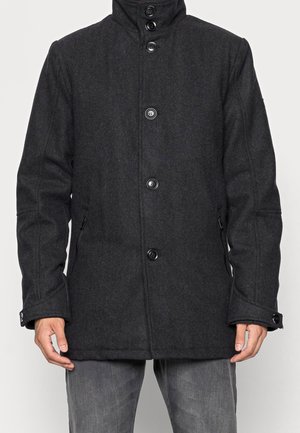 Homme portant un manteau en laine gris foncé boutonné avec des poches latérales zippées et des poignets boutonnés, debout devant un fond uni.