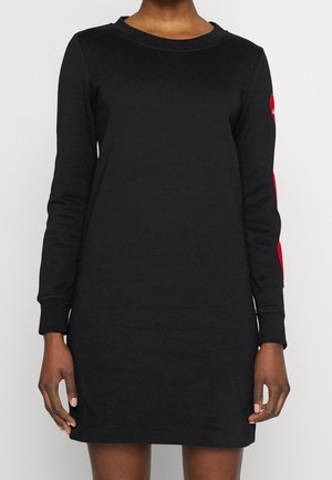 Femme portant une robe mini noire à manches longues avec des patchs rouges sur la manche gauche, debout devant un fond blanc uni.