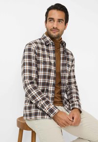Camicia a quadri marrone, bianca e blu navy con bottoni sul davanti; maniche lunghe, due tasche sul petto, indossata sopra un dolcevita marrone, seduta su uno sgabello.
