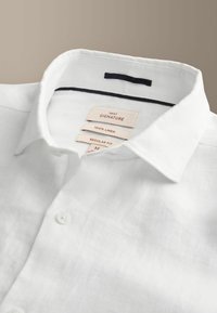 Chemise en lin blanc avec col italien, boutons et étiquettes indiquant "Next Signature", "100 % lin" et "Regular Fit, M" à l'intérieur du col.