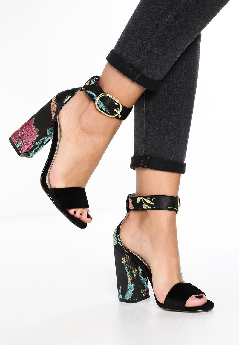 Pieds portant des sandales noires à talons hauts avec des motifs floraux sur des talons épais et des brides autour de la cheville, associés à un jean skinny foncé retroussé.