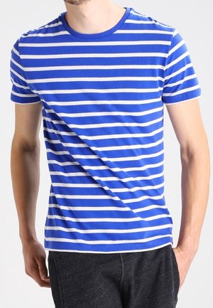 Mann trägt ein blau-weiß gestreiftes T-Shirt mit kurzen Ärmeln und dunkle Hose, steht mit einer Hand in der Tasche.