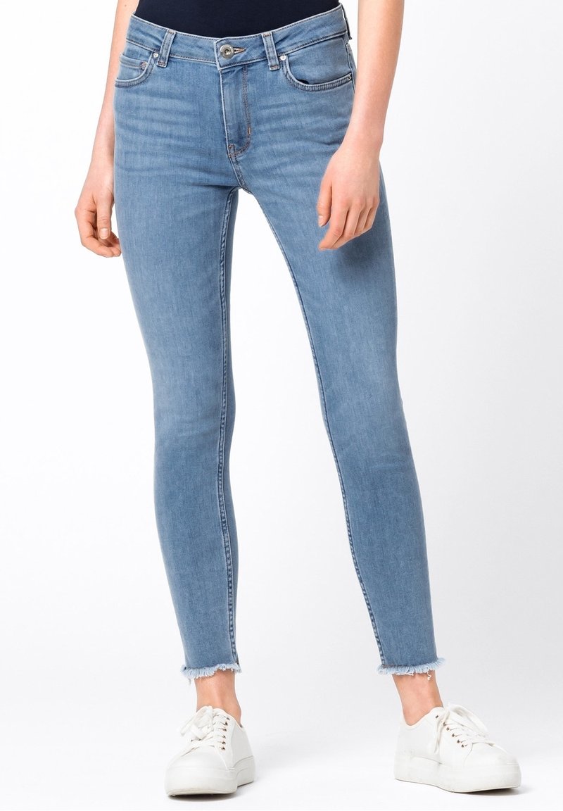 Slim fit ljusblå denimjeans med fransade fållar, bärs med vita sneakers och en mörk topp, visad på en stående person.