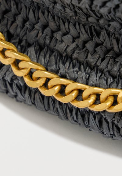 Κοντινή λήψη μαύρου υφάσματος με πλεκτή υφή και λεπτομέρεια χοντρής χρυσής αλυσίδας που ακουμπά στην επιφάνειά του πάνω σε λευκό φόντο.
