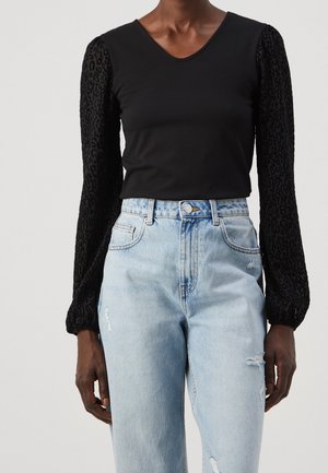 Femme portant un haut noir à manches longues avec des manches transparentes à motif léopard et un jean taille haute bleu clair déchiré.