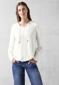 Felpa bianca leggera con scollo a V e coulisse; presenta maniche lunghe e un tessuto strutturato. Abbinata a jeans in denim blu.