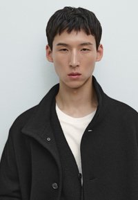 Cappotto nero in misto lana con grande colletto e dettagli con bottoni, sovrapposto a una t-shirt bianca. Texture liscia e design strutturato.