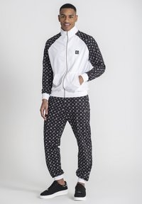 Chándal con chaqueta blanca de cremallera y mangas de patrón negro y blanco, combinado con pantalones negros a juego y zapatillas negras.
