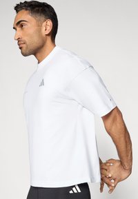 T-shirt branca de manga curta feita de algodão macio. Apresenta decote redondo e um emblema cinza no peito. Design limpo e minimalista.