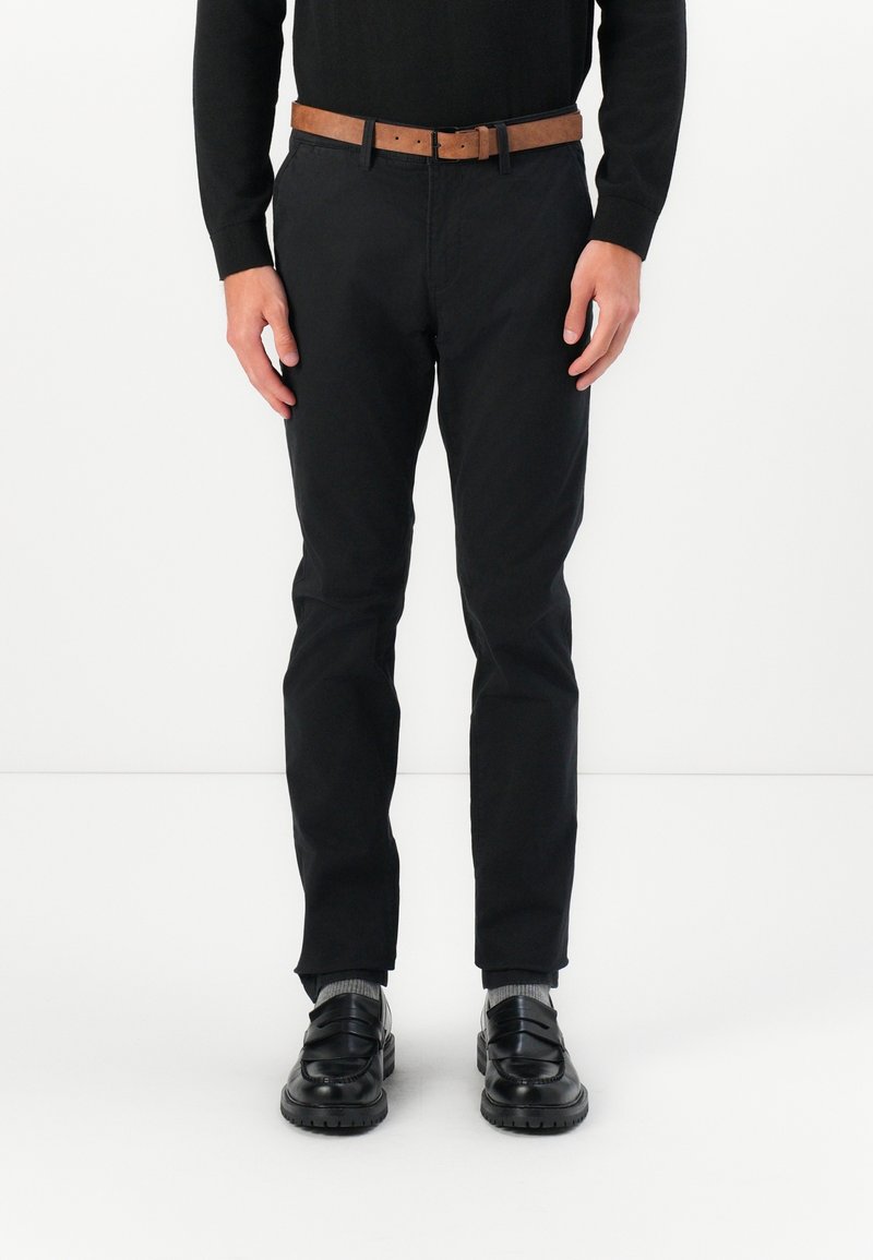 Schwarze Hose mit geradem Schnitt, getragen mit einem braunen Ledergürtel und schwarzen Slippern. Graue Socken sind über den Schuhen sichtbar. Weißer Hintergrund.