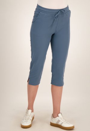 Blauwe cropped broek met zijsplits gedragen met witte sneakers bij een staand persoon tegen een neutrale achtergrond.