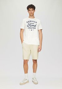 Camiseta blanca de manga corta con estampado azul "SERVICE Ford CENTER", combinada con pantalones cortos beige, calcetines blancos y zapatillas ligeras.