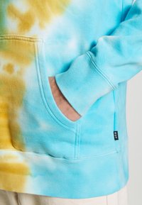 Sudadera de tie-dye azul claro con un bolsillo frontal, puños acanalados y una pequeña etiqueta en el costado. Tejido suave con un acabado texturizado.