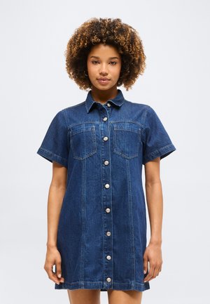 Νεαρή γυναίκα με σγουρά μαλλιά, που φοράει σκούρο τζιν φόρεμα με κοντά μανίκια και κουμπιά μπροστά, στέκεται μπροστά σε ένα απλό φόντο.