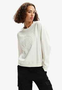 Sudadera blanca con un logo texturizado en el centro. Presenta un cuello redondo y mangas largas con puños. Combinada con pantalones cargo negros.